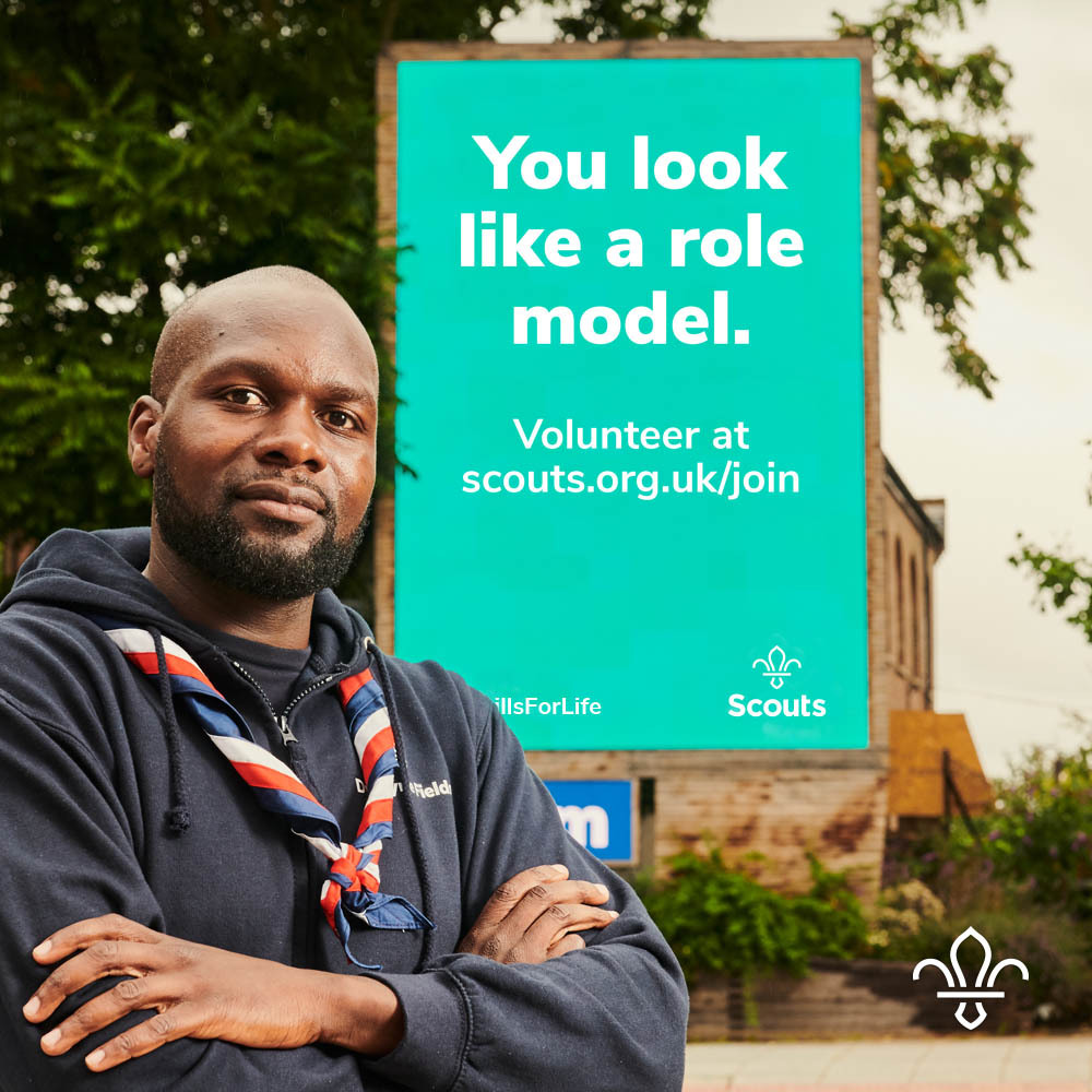 Chief Scout, Dwayne Fields, stands in front of a billboard reading "You look like a role model. Volunteer at scouts.org.uk/join"